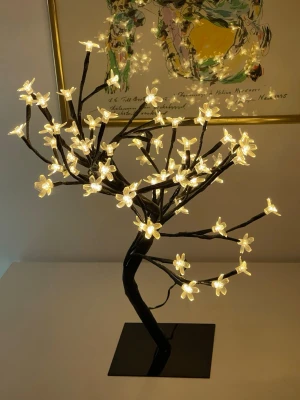 Dekorativ LED blomsterlampa - Säljer en dekorativ LED-lampa formad som ett körsbärsträd med genomskinliga blommor som lyser i varmt vitt sken. Lampan har svarta grenar och står på en fyrkantig svart fot. Perfekt som mysig belysning i rummet eller som snygg inredningsdetalj.