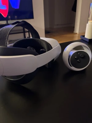 Meta Quest 2 VR-headset med handkontroller - Säljer ett Meta Quest 2 VR-headset i vit plast med matchande handkontroller. Headsetet har justerbart svart band och bekväm vaddering runt ögonen. Perfekt för gaming och VR-upplevelser hemma. Snygg och modern design som passar alla gamers.