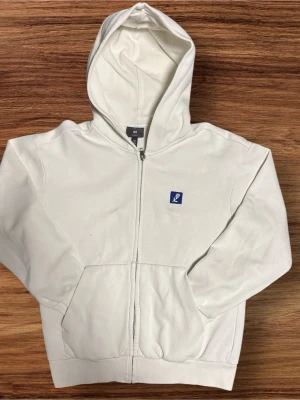 Vit zip hoodie från H&M - Vit hoodie med dragkedja från H&M. Klassisk modell med huva och känguruficka framtill. Liten blå patch med logga på bröstet. Tillverkad i mjukt bomullsmaterial, perfekt för en avslappnad stil.