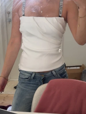 Vit axelbandslös topp med drapering - Snygg vit axelbandslös topp med draperad framsida och raka linjer. Toppen har breda axelband med logga och sitter tight mot kroppen. Perfekt till jeans eller kjol för en trendig look.