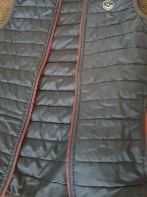 Svart dunväst från Jack & Jones - Snygg svart dunväst från Jack & Jones med horisontella sömmar och röda dragkedjedetaljer. Västen har en klassisk, quiltad design och är lätt vadderad för extra värme. Perfekt att slänga över en hoodie eller tröja för en cool lager-på-lager-look.