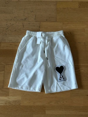 Vita shorts från Ami Paris med hjärtdetalj - Säljer ett par vita shorts från Ami Paris med elastisk midja och snörning. På vänster ben finns en stor svart hjärtdetalj med bokstaven A. Shortsen har sidofickor och är tillverkade i mjuk bomull, perfekta för sommardagar. Det står storlek M men passformen är som storlek S.