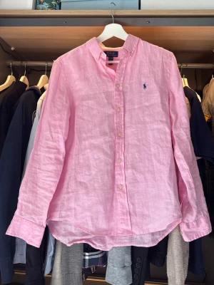 Rosa linneskjorta från Polo Ralph Lauren - Säljer en snygg rosa skjorta i linne från Polo Ralph Lauren. Skjortan har klassisk krage, knappar framtill och lång ärm. Ikoniska loggan broderad på bröstet. Perfekt för en fräsch och stilren look. OBS DEN HAR ETT LITET LITET HÅL DÄR EN TRÅD HAR GÅTT AV. Går att fixa enkelt. Det syns inte enligt mig. Där av är den så billig.😁🤝