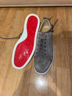 Gråa sneakers från Christian Louboutin - Snygga gråa sneakers i mocka från Christian Louboutin med klassisk röd sula och vita kanter. Skorna har rund tå, matchande grå skosnören och diskreta sömmar för en clean look. Perfekta för dig som gillar exklusiva detaljer och streetstyle.