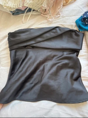 Svart satin tubtopp med omlott - Snygg svart axelbandslös tubtopp i satin med omlottdetalj upptill. Toppen har en elegant glansig finish och en lätt utsvängd passform. Perfekt för att skapa en trendig och stilren look.