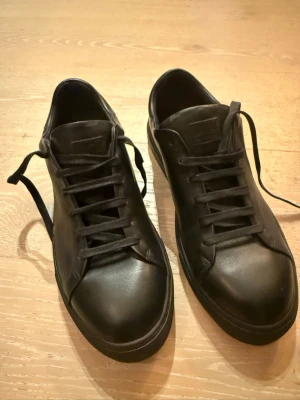 Svarta sneakers från J.Lindeberg - Nya - Stilrena svarta sneakers från J.Lindeberg i slätt läder med matchande svarta skosnören och diskret logga på plösen. Klassisk låg modell som passar till det mesta och ger en clean look. Perfekta för dig som gillar minimalistisk och tidlös stil. Skorna är helt nya!