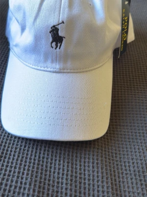 Vit keps från Polo Ralph Lauren - Snygg vit keps från Polo Ralph Lauren med klassisk broderad polospelare framtill och 'POLO' broderat baktill. Justerbar rem med metallspänne där det också står Polo. Tillverkad i bomull för en skön och luftig känsla.