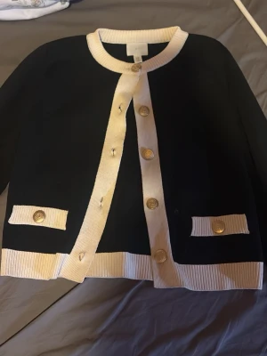 Svart kofta med vita detaljer H&M XS - Stilren svart kofta från H&M i storlek XS med vita ribbade kanter och guldknappar. Koftan har rund halsringning och två dekorativa fickor framtill. Perfekt för en chic och klassisk look.