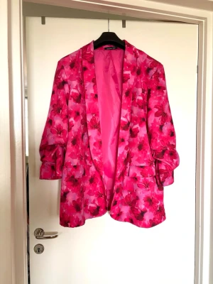 Blommig rosa kavaj med mönster plussize - Snygg kavaj i starkt rosa med blommigt mönster. Oversized passform med breda slag och två fickor framtill. Perfekt statement-plagg för dig som vill sticka ut. Tunnare material med blank finish och helfodrad insida. Aldrig använd. 