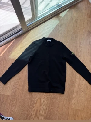 Svart stickad tröja från Stone Island - Snygg svart stickad tröja från Stone Island med rund halsringning och klassisk logotyp-patch på vänster ärm. Tröjan har en stilren och enkel design med långa ärmar och ribbade muddar. Perfekt för dig som gillar streetwear och exklusiva märken.