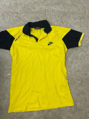 Gul och svart pikétröja från Nike - Nike pikétröja i gul med svarta detaljer på krage och ärmar. Klassisk Nike-logga på bröstet och 'Nike Air' tryckt på axeln. Skön passform och perfekt för en sportig look.