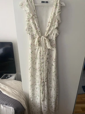 Vit långklänning med volang och knyt - Långklänning i vit med blommigt mönster och volangdetaljer längs axlar och urringning. Djup v-ringning framtill med knyt i midjan. Ärmlös design och lätt, flödande material som passar perfekt till sommaren🌸🌸