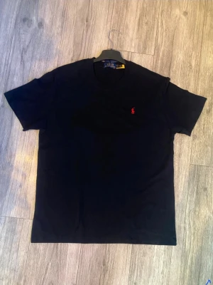 Svart t-shirt från Polo Ralph Lauren - Svart t-shirt från Polo Ralph Lauren med klassisk passform och rund hals. T-shirten har en liten röd broderad logga på bröstet och är tillverkad i mjuk bomull. Perfekt för en clean och stilren look. PRISLAPP FINNS MEN SYNS INTE PÅ BILDEN, DUSTBAG RAPH LAUREN FINNS OCKSÅ. FRÅGOR PÅ DETTA, DE E BARA SKRIVA. SKICKAR BILDER!!