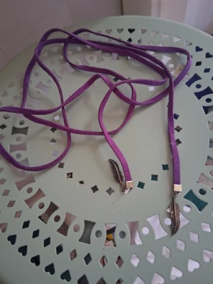 Lila halsband med fjäderhängen - Snyggt lila halsband i mockaimitation med två silverfärgade fjäderhängen i metall. Halsbandet är långt och kan stylas på olika sätt för en personlig look. Perfekt accessoar för att lyfta din outfit.