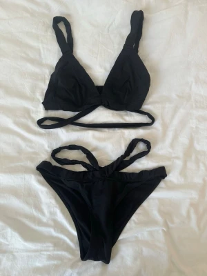 Svart bikini med twistade band - Snygg svart bikini med twistade axelband och extra banddetaljer både upptill och nedtill. Bikinin har en enkel men trendig design med triangelformad överdel och högt skurna trosor. Perfekt för stranden eller poolen. ÖVERDELEN ÄR I STORLEK 42 OCH UNDERDELEN I STORLEK 38