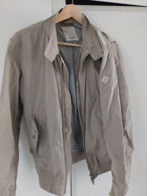 Beige vindjacka från J.Lindeberg - Stilren beige vindjacka från J.Lindeberg med klassisk krage, dragkedja och tryckknappar vid fickorna. Jackan har en diskret logotyp på ärmen och är tillverkad i ett lätt, vindtätt material. Perfekt för en snygg och avslappnad look. Något missfärgad vid nacken
