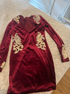 Vinröd sammetsklänning från NLY One - Säljer en vinröd sammetsklänning från NLY One med guldiga broderade detaljer och djup v-ringning. Klänningen har lång ärm och figurnära passform, perfekt för dig som vill sticka ut. Dragkedja i ryggen för enkel påklädning.
