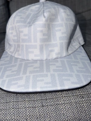 Vit keps med Fendi-logga - Snygg vit keps från Fendi med det klassiska FF-mönstret över hela kepsen. Kepsen har rak skärm och är tillverkad i ett tygmaterial som känns både lyxigt och trendigt. Perfekt accessoar för att lyfta din outfit.