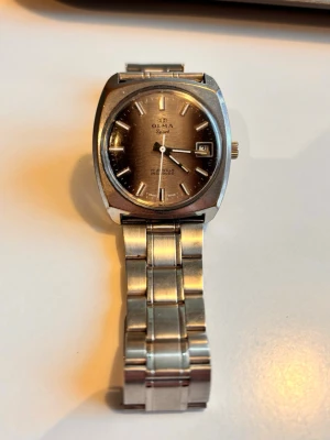 Olma Signet herrklocka i silver - Snygg herrklocka från Olma, modell Signet, med klassisk rund urtavla i bronsfärg och datumvisning. Klockan har ett robust armband i silverfärgad metall och stilrena indexmarkeringar. Perfekt accessoar för dig som gillar vintage och tidlös design.