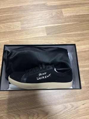 Svarta Saint Laurent sneakers - Svarta sneakers från Saint Laurent med vit logga på sidan och beige sula. Skorna har klassisk låg profil, svarta skosnören och ovandel i canvas. Pris kan diskuteras 