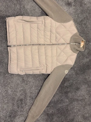 Moncler cardigan  - Snygg beige cardiganjacka med quiltad framsida och stickade ärmar samt rygg. Dragkedja framtill och två stora fickor. Hög krage och ribbade muddar för extra komfort. Perfekt för lager-på-lager och en clean look.
