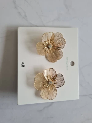 Guldfärgade blomörhängen H&M - Stora örhängen i guldfärgad metall från H&M, formade som blommor med tunna, räfflade kronblad och en liten glittrande sten i mitten. Perfekta för dig som vill ha ett statement-smycke med elegant touch.