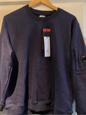 Mörkblå sweatshirt från C.P. Company - Säljer en mörkblå sweatshirt från C.P. Company. Tröjan har den ikoniska linsen på ärmen. Perfekt för sommarn då den är tunn men kan även användas under vår/höst. Tröjan är storlek L men är liten i storleken. Tröjan är aldrig använd. Priset kan diskuteras vid en snabb affär🤝✅