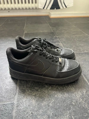 Svarta Nike Air Force 1 sneakers - Klassiska helsvarta Nike Air Force 1 sneakers i lågt utförande. Skorna har ovandel i läder, perforerad tåbox och svart sula. Swoosh-logga på sidan och metallbricka på snörningen. Perfekta för en clean och stilren look.