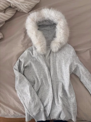 Grå hoodie med fuskpäls från Hollister - Mysig grå hoodie från Hollister med stor huva kantad av vit fuskpäls. Tröjan har två fickor framtill och ribbade muddar vid ärmslut och nederkant. Älskar den men för liten på mig tyvärr. Aldrig använd