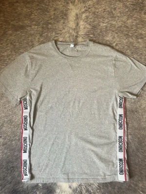 Grå Moschino t-shirt med logoband - Snygg grå t-shirt från Moschino med vita logoband längs sidorna där det står MOSCHINO i svart text och en röd linje. Klassisk rund hals och korta ärmar. Perfekt för dig som gillar streetwear och vill ha något stilrent men ändå med en twist.