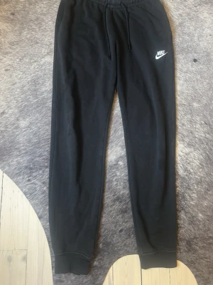 Svarta mjukisbyxor från Nike - Svarta mjukisbyxor från Nike med klassisk vit logga på vänster lår. Byxorna har dragsko i midjan och muddar vid bensluten. Perfekta för en avslappnad stil och tillverkade i mjukt material för extra komfort.
