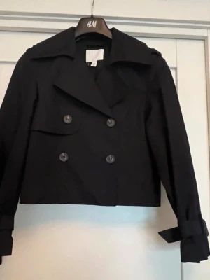 Kort svart trenchcoat från H&M - Stilren svart trenchcoat från H&M med dubbelknäppning framtill, bred krage och knytdetaljer vid ärmsluten. Jackan är kortare i modellen och har en klassisk design som passar perfekt till många olika outfits. Tillverkad i ett slätt tyg.