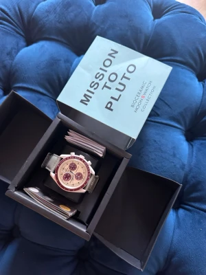 Swatch x Omega Mission to Pluto klocka - Cool klocka från Swatch x Omega Bioceramic MoonSwatch Collection, modell Mission to Pluto. Klockan har en ljusgrå boett, vinröda detaljer på urtavlan och svart armband. Snygg och unik design med kronograf och tydliga index. Levereras i originalbox.