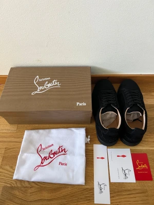 Christian Louboutin svarta sneakers - Snygga svarta sneakers från Christian Louboutin med klassisk röd sula. Skorna har lågt skaft, svarta skosnören och är tillverkade i mocka. Kommer med originalkartong, dustbag och tillbehör. Perfekta för dig som vill sticka ut med exklusiv stil.