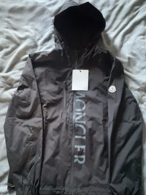 Svart Moncler vindjacka med huva - Svart vindjacka från Moncler med stor logotyp längs dragkedjan och klassisk Moncler-patch på ärmen. Jackan har huva, bröstficka med dragkedja och elastiska muddar. Materialet är tunt och vattenavvisande, perfekt för blåsiga dagar.
