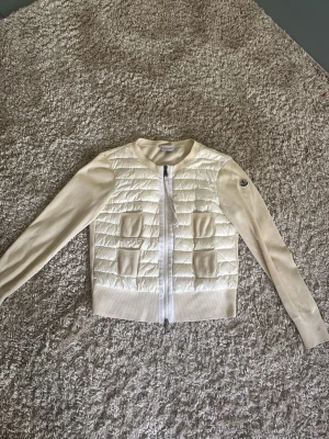 Moncler cardigan - Snygg vit cardiganjacka från Moncler med quiltad framsida och två fickor. Jackan har ribbade muddar, dragkedja framtill och Moncler-logga på ärmen. Ärmarna är stickade medan framsidan är i glansigt material. Perfekt för lager på lager och en clean look. 