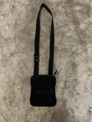 Svart axelväska från BOSS - Snygg svart axelväska från BOSS med justerbart axelband och diskret logga på framsidan. Väskan har en minimalistisk design och är tillverkad i ett slittåligt material med dragkedja upptill. Perfekt för att bära det viktigaste med stil.