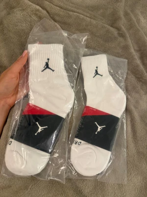 Vita Jordan strumpor med logga - Säljer ett par vita Jordan strumpor med svart Jumpman-logga på sidan. Strumporna är ribbade upptill och tillverkade i en bomullsblandning för extra komfort. Perfekta för dig som gillar sportig stil och vill ha snygga detaljer på dina strumpor. Storlek 8-12 Eu