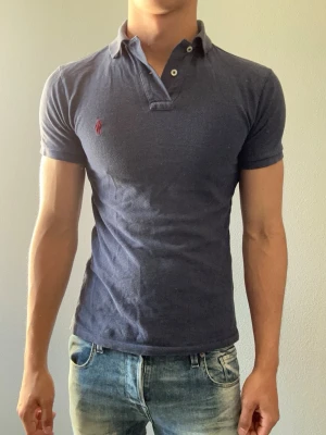 Mörkblå pikétröja från Ralph Lauren - Klassisk mörkblå pikétröja från Ralph Lauren med röd broderad logga på bröstet. Tröjan har krage, korta ärmar och knappar vid halsen. Tillverkad i mjuk bomull och har en slim passform som sitter snyggt på kroppen.