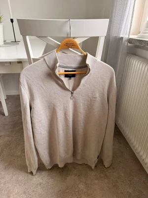 Beige halvzip-tröja från bondelid - Snygg beige tröja från Bondelid med halvzip och ribbad krage. Tröjan har långa ärmar och diskret broderad logga på bröstet. Tillverkad i mjukt material som passar perfekt till lager-på-lager-stil.