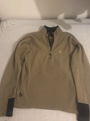 Beige fleece från Haglöfs med halvzip - Säljer en beige fleece från Haglöfs med halvzip och hög krage. Tröjan har svarta detaljer vid ärmslut och krage samt en liten Haglöfs-logga på bröstet.
