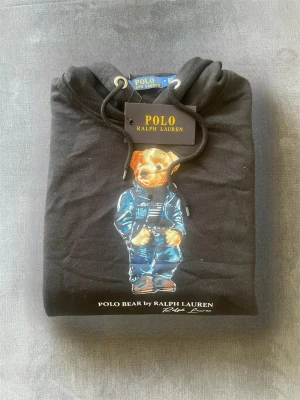 Svart hoodie Polo Bear Ralph Lauren - Svart hoodie från Polo Ralph Lauren med ikoniskt Polo Bear-motiv på bröstet. Klassisk huva med dragsko och tryckt logga under björnen. Perfekt för en avslappnad och trendig stil.