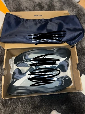 CD blå och vita sneakers - Snygga sneakers från Asics i blått, vitt och svart med mesh- och syntetmaterial. Skorna har vita snören och en chunky sula som ger en sportig vibe. Perfekta för dig som gillar streetwear och vill ha bekväma skor med stil.