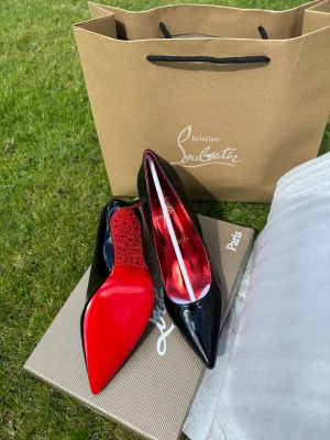 Svarta klackar från Christian Louboutin - Snygga svarta klackskor från Christian Louboutin med klassisk röd sula och röda glittriga detaljer på hälen. Skorna har spetsig tå och är tillverkade i lackat material som ger en lyxig känsla. Skorna är aldrig använda och i topp skick, skriv t mig för mer kvalitet bilder dessa på plick blir suddiga