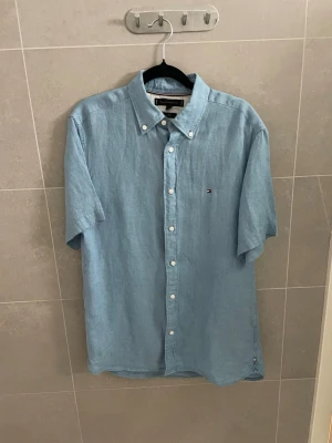 Blå kortärmad linneskjorta från Tommy Hilfiger - Helt ny! Fick den i present men den passade inte. Snygg ljusblå skjorta i linne från Tommy Hilfiger med korta ärmar och klassisk button-down krage. Skjortan har vita knappar och en liten broderad logga på bröstet. Perfekt för en avslappnad och stilren look. skrivas ifall bra bilder krävs. Lite skrinkliga o så men det försvinner i tvätten eller om du ska strika dom eller hur du än gör.