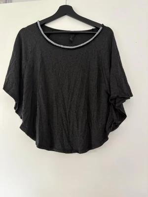 Svart loose topp med silverdetalj - Snygg svart topp med lös passform och korta, vida ärmar. Rund halsringning med en silverfärgad detalj som ger en cool touch. Perfekt till jeans eller kjol för en avslappnad look. Storlek M