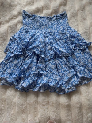 Blå volangkjol  - Supersöt kort kjol med blått och vitt blommönster och flera volanger. Kjolen har smock i midjan för skön passform och är gjord i ett lätt, luftigt material. Perfekt för dig som vill ha en feminin och trendig look.