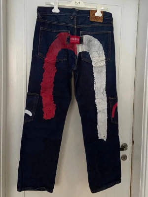 Evisu jeans intressekoll - Unika mörkblå jeans från Evisu med handmålat rött och vitt mönster på bak- och framsida. Klassisk femficksmodell i kraftig denim med raka ben och stora sidfickor. Perfekta för dig som vill sticka ut med streetwear-inspirerad stil.                                                ———-                                                         Hade ett hål i skrevet som jag lagat, lappen får bak lossnade på ena sidan men sydde tillbaka det        Bara säg till om du har frågar eller fler bilder