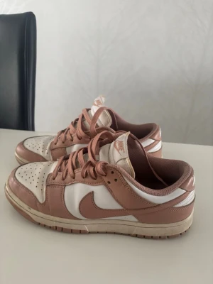 Nike Dunk Low rosa och vita sneakers - Nike Dunk Low sneakers i en snygg kombo av ljusrosa och vitt. Skorna har klassisk låg siluett, perforerad tå och Nike-logga på sidan. Ovandelen är i läder med matchande rosa snören och vit sula. Perfekta för dig som gillar streetstyle och vill sticka ut.