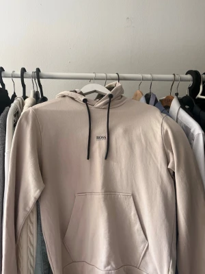 Beige hoodie från BOSS med huva - Snygg beige hoodie från Hugo BOSS med svart dragsko i huvan och liten Hugo BOSS-logga på bröstet. Hoodien har en klassisk känguruficka framtill och mönstrat innerfoder i huvan. Perfekt för en chill och stilren look.